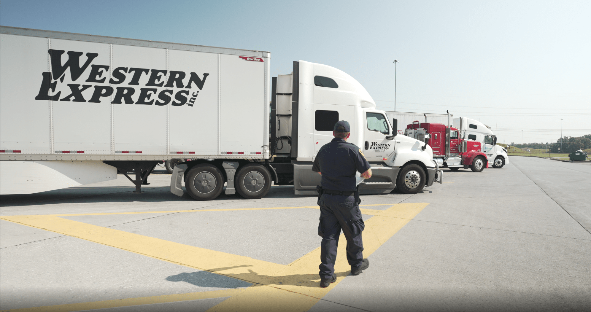 A man in uniform strides through a paved lot lined with semi-trucks, including one marked Western Express. Under the clear sky, he represents the essence of trucking and partnerships that keep commerce moving smoothly.