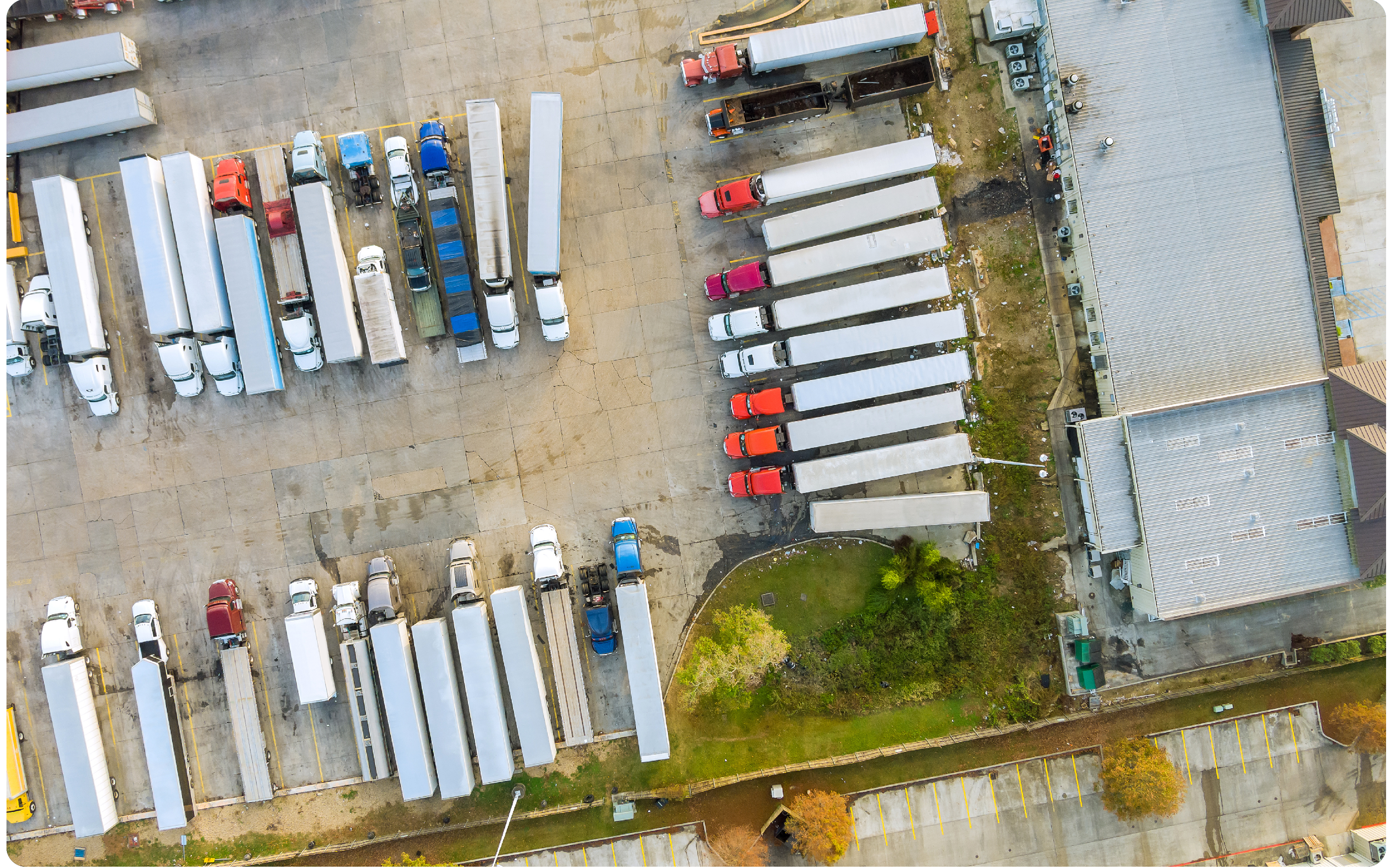 Aerial view of a warehouse parking lot with rows of semi-trucks and trailers, some equipped with ELD devices. Trucks are positioned near loading docks on the right. Sparse greenery and a small road run along the bottom, ensuring smooth preclear processes for logistics partners.