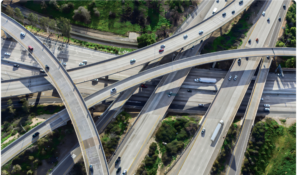 Aerial view of a complex freeway interchange with multiple overlapping highways, surrounded by greenery. Various trucks and vehicles travel the roads, illustrating busy traffic patterns essential for trucking. The image highlights the intricacies of urban infrastructure.