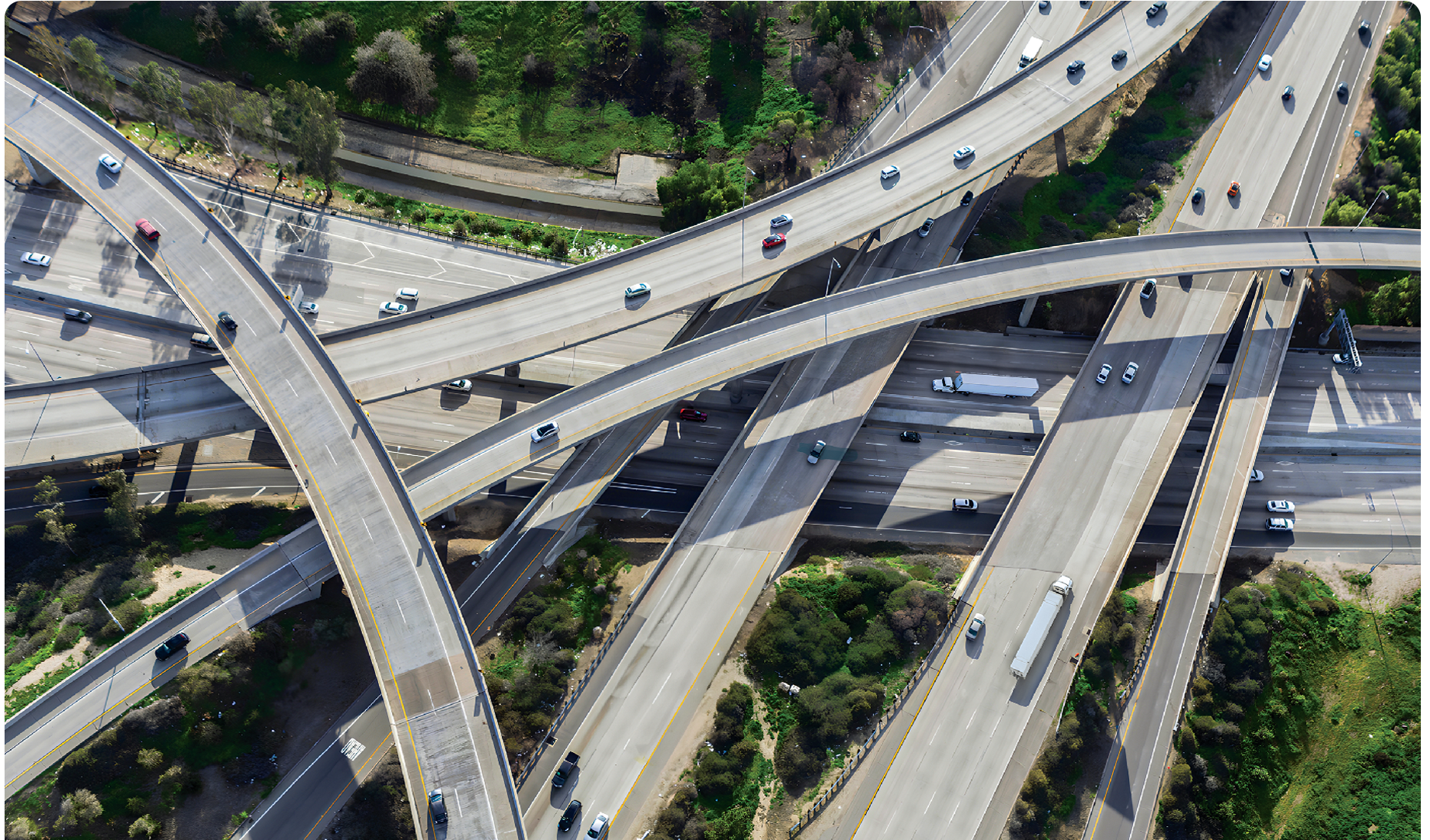 Aerial view of a complex freeway interchange with multiple overlapping highways, surrounded by greenery. Various trucks and vehicles travel the roads, illustrating busy traffic patterns essential for trucking. The image highlights the intricacies of urban infrastructure.