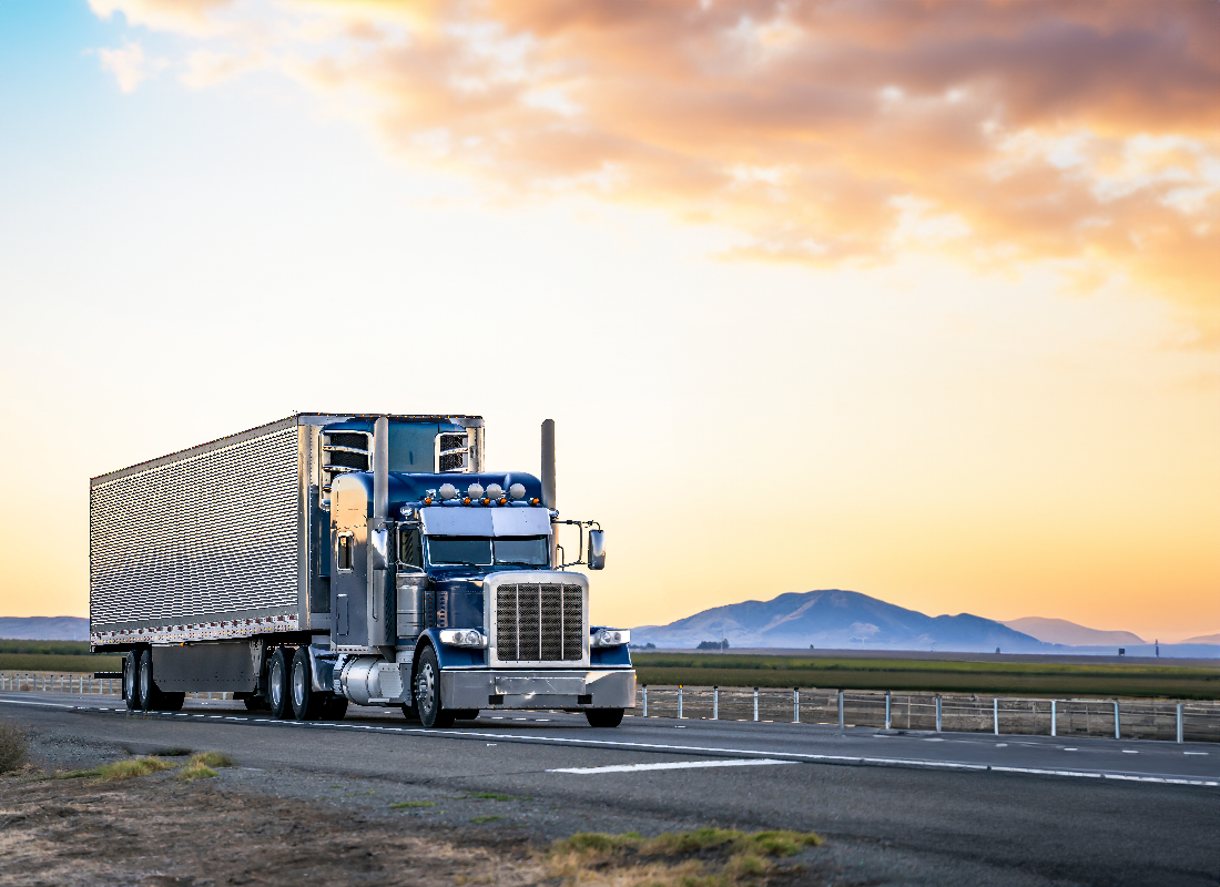 A blue semi-truck with a large trailer, unmistakably belonging to a seasoned trucker, drives down a highway during sunset. The sky is a stunning mix of orange and purple hues, with mountains in the background. The empty road exudes a sense of journey and the freedom of the open road.