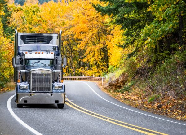 A large semi-truck, equipped with the latest ELD technology, drives down a winding two-lane road surrounded by vibrant autumn trees with yellow and green leaves. The road has double yellow lines, and scattered leaves are visible on the ground.