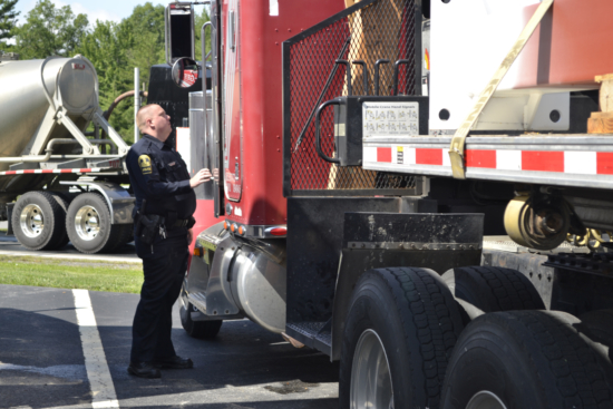 A police officer inspects a red semi-truck parked in a lot, in collaboration with their partner. The officer, wearing a crisp uniform, stands by the trucks cabin while another tanker truck is visible parallel to the semi. Trees stretch into the distance, framing this roadside preclear scene.