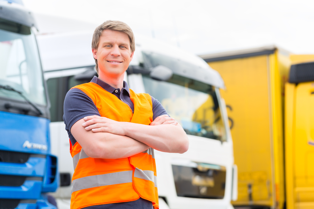 A man wearing an orange safety vest stands with arms crossed and smiles in front of several parked trucks, confident in his fleet’s strong CSA score, with blue, white, and yellow vehicles visible behind him.