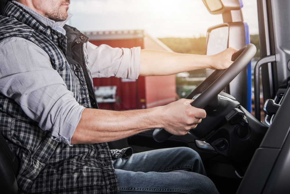 A man wearing a plaid vest and rolled-up shirt sleeves sits in the driver’s seat of a truck, holding the steering wheel with both hands, demonstrating fleet safety. A red shipping container is visible outside the window.