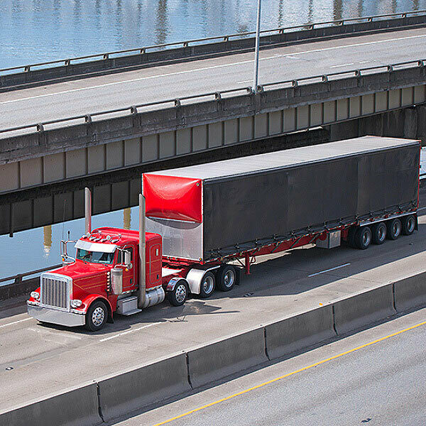 A red semi-truck with a black trailer drives on a highway next to a concrete barrier, with an overpass and water visible in the background.