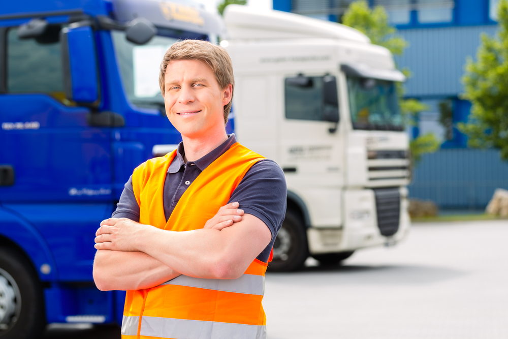A smiling man wearing an orange safety vest stands with arms crossed in front of two parked trucks, showcasing Drivewyze Weigh Station Bypass in an outdoor industrial or transport setting.