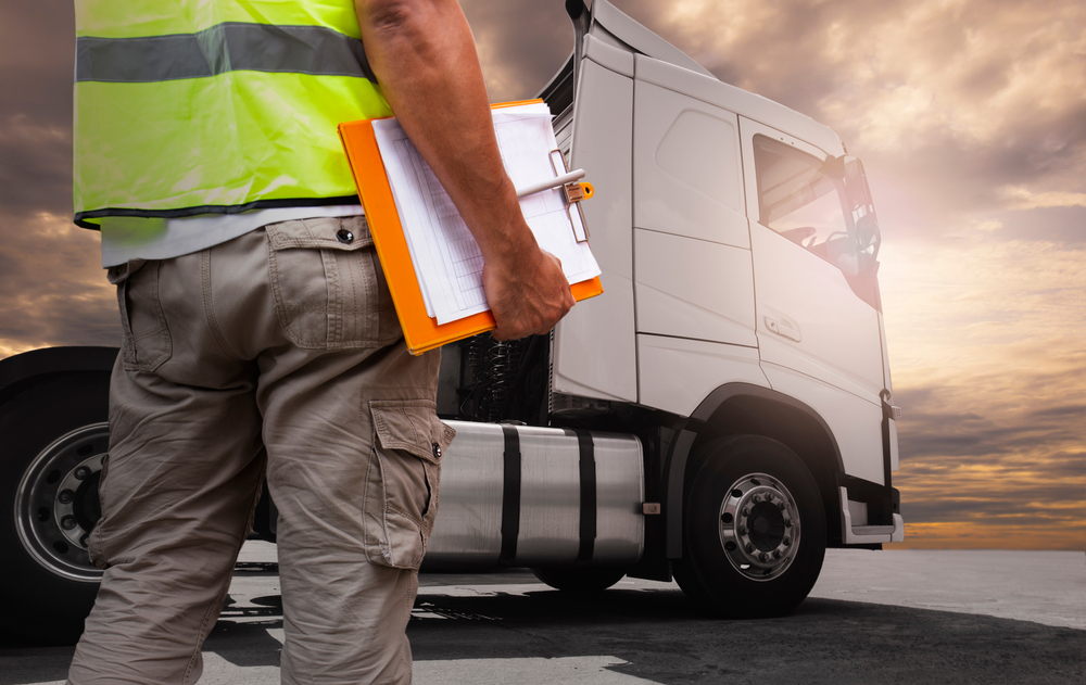 A person in a yellow safety vest holding a clipboard stands near a white semi-truck under a dramatic, cloudy sky at sunset, perhaps preparing to check how many hours a day a trucker can drive.