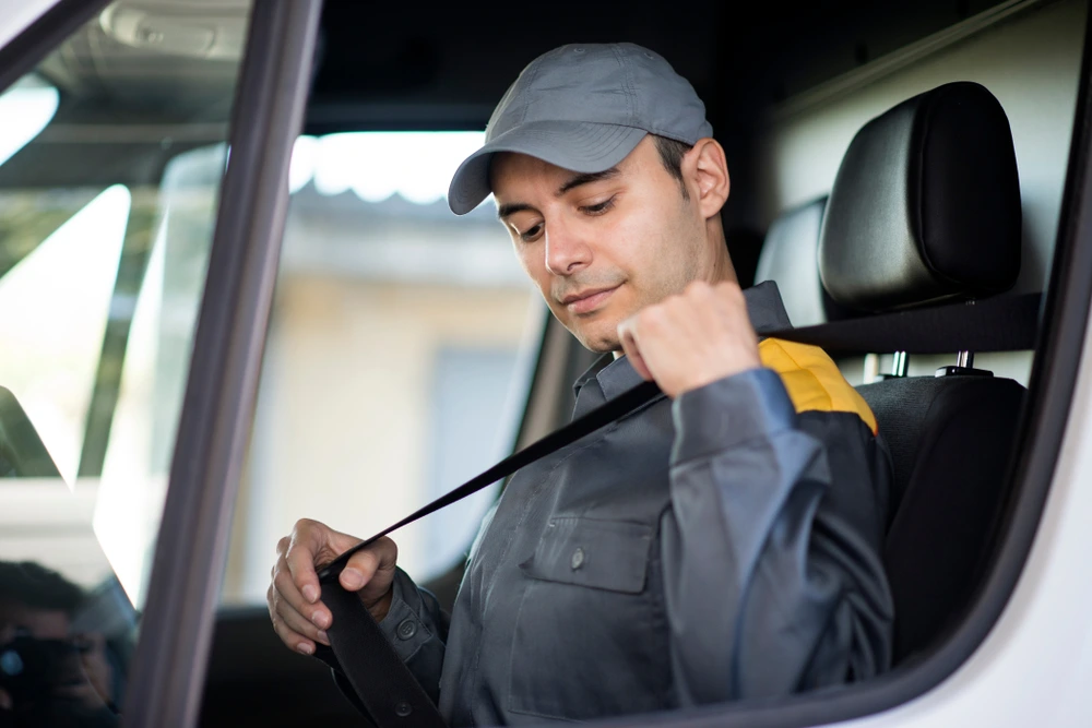 A delivery driver in a grey uniform and cap sits in a vehicle and fastens his seatbelt, preparing for a drive—an essential first step for those wondering how to get good gas mileage in a truck.