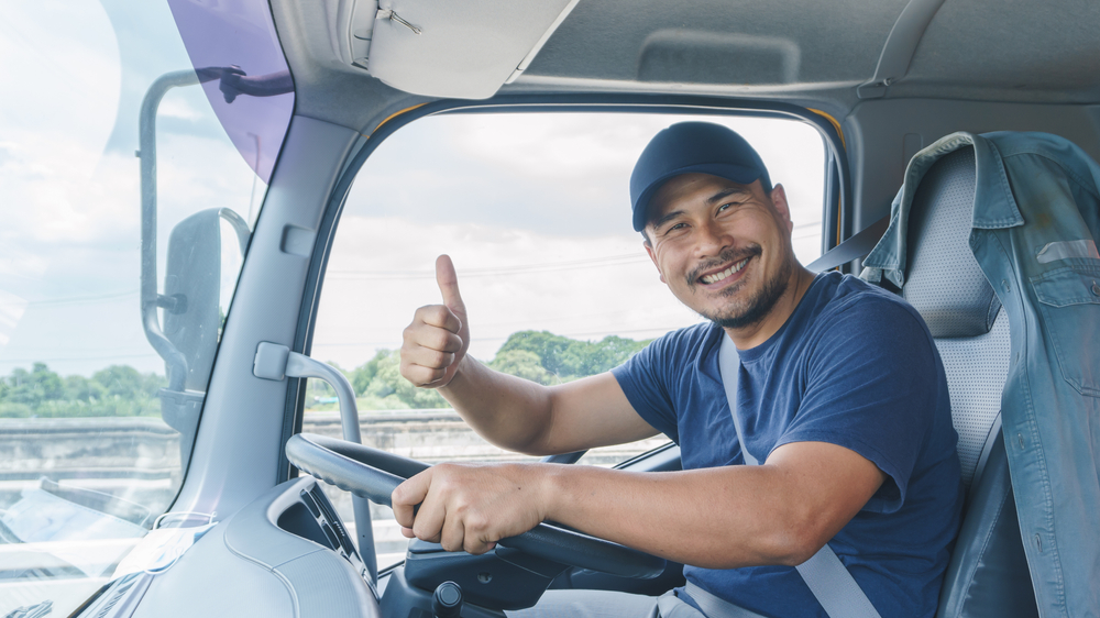 A smiling man wearing a cap and seatbelt sits in the truck’s driver’s seat, giving a thumbs-up—perhaps after checking how many hours a day a trucker can drive—with trees and sky visible through the window.